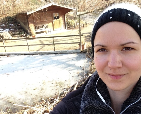 Eine junge Frau steht an einem zugefrorenen Teich. Sie trägt eine schwarz-weiße Bommelmütze und einen fluffigen Sweater.