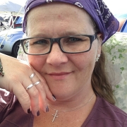 Eine Frau mit einer schwarzen, eckigen Brille und schulterlangen, hellbraunen Haaren. sie trägt ein lilafarbenes Kopftuch und ein lila Shirt.