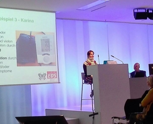 Eine Frau mit kurzen, braunen Haaren und einem 50er Jahre Kleid sitzt hinter einem Podium und gibt einen Vortrag zum Thema Ehlers-Danlos-Syndrom