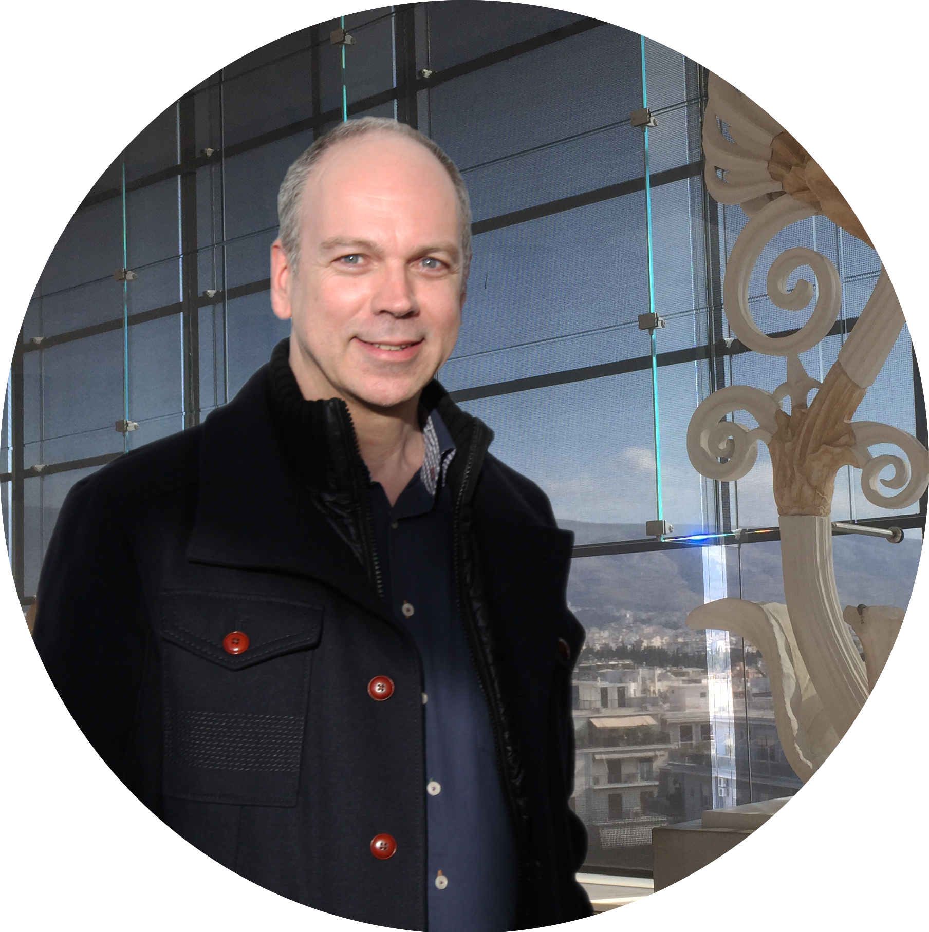 This image shows Uwe Strauch (CEO German Museums Magazin) who wears a black jacket with red buttons, a dark blue shirt and stands in front of a sculpture. He has blue eyes and very short, grey hair.