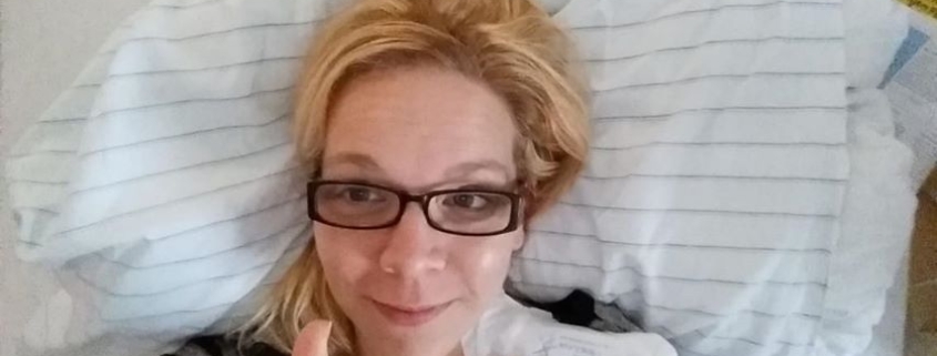 Eine junge Frau mit blonden Haaren und einer eckigen, schwarzen Brille liegt in ihrem Krankenhausbett und hat einen Katheter an der linken Halsseite.