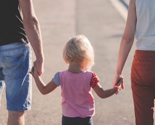 Auf dem Bild zu sehen sind ein Elternpaar und ein Kind von hinten. Links ist ein Mann in blauen, kurzen Jeanshosen, das ein Kind mit kurzen blonden Haaren und rosa T-Shirt an der Hand hält. Rechts von dem kleinen Mädchen ist eine Frau mit roten Hosen.