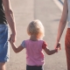 Auf dem Bild zu sehen sind ein Elternpaar und ein Kind von hinten. Links ist ein Mann in blauen, kurzen Jeanshosen, das ein Kind mit kurzen blonden Haaren und rosa T-Shirt an der Hand hält. Rechts von dem kleinen Mädchen ist eine Frau mit roten Hosen.
