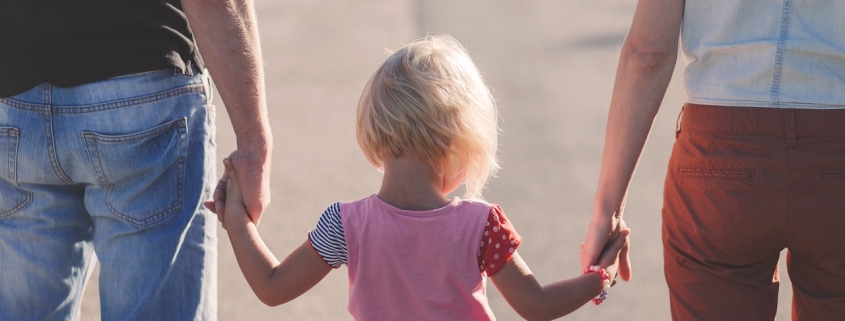 Auf dem Bild zu sehen sind ein Elternpaar und ein Kind von hinten. Links ist ein Mann in blauen, kurzen Jeanshosen, das ein Kind mit kurzen blonden Haaren und rosa T-Shirt an der Hand hält. Rechts von dem kleinen Mädchen ist eine Frau mit roten Hosen.