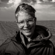 Schwarz-weiß Porträtaufnahme einer Frau mit kurzen blonden Haaren, die im Wind wehen. Sie lächelt, trägt eine Brille mit breitem, durchsichtigen Rahmen und eine schwarze Jacke.