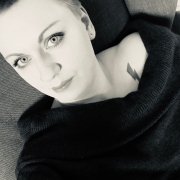Schwarz-weiß Aufnahme einer jungen Frau mit kurzen Haaren. Sie sitzt auf einem Sofa, trägt einen schwarzen Pullover mit u-förmigen Ausschnitt in dem man ein Tattoo eines Blitzes erkennt.