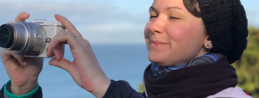 Eine Frau mit Beanie und Halskrause macht Bilder von Fort Funston in San Francisco und lächelt dabei glücklich.