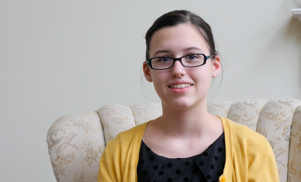 Mid-size frontal shot of a girl with brown hair bound to a ponytail. She wears black glasses and a yellow cardigan.