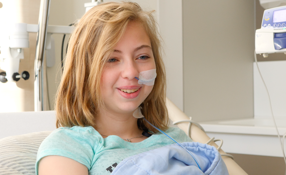 Mid-size shot of a girl with shoulder-long blonde hair and blue eyes. She is in a hospital bed and has a tube in her nose.
