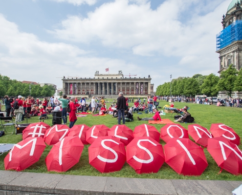 Viele Menschen sitzen auf einer Wiese und tragen rote Shirts. Auf mehreren Regenschirmen sind Buchstaben abgedruckt, die die Worte Millions Missing ergeben. Protestveranstaltung in Berlin.