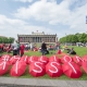 Viele Menschen sitzen auf einer Wiese und tragen rote Shirts. Auf mehreren Regenschirmen sind Buchstaben abgedruckt, die die Worte Millions Missing ergeben. Protestveranstaltung in Berlin.