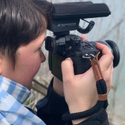 Side view of a woman with short, brown hair holding a camera with a big microphone in front of her face and filming out to the ocean.