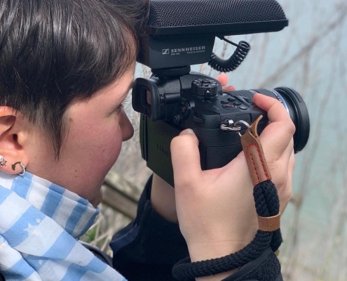 Side view of a woman with short, brown hair holding a camera with a big microphone in front of her face and filming out to the ocean.