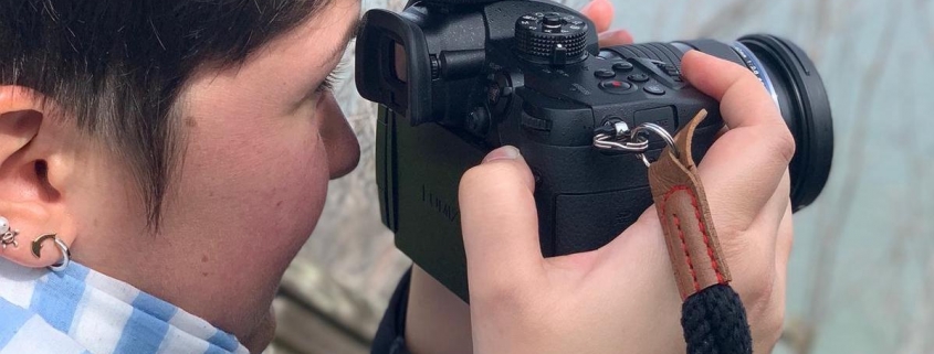 Side view of a woman with short, brown hair holding a camera with a big microphone in front of her face and filming out to the ocean.