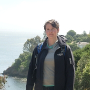 Eine Frau mit kurzen braunen Haaren und einer Blume im Haar steht vor einer Klippe auf Angel Island. Hinter ihr ist eine Landzunge mit vielen grünen Bäumen.