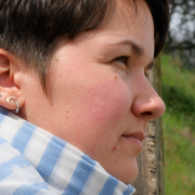 Karina, a woman with short, brown hair looks into the nothingness. She sits on stairs in the nature and is seen in profile. She wears four earrings. One of those is a dolphin, and the other one a turtle. Additionally, she wears a white-blue scarf around her neck brace.