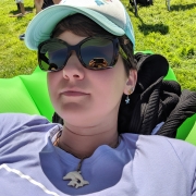 Eine Frau mit kurzen braunen Haaren liegt auf einer grünen aufblasbaren Couch im Grünen. Sie trägt eine Türkise Mütze und Sonnenbrille.