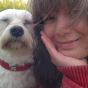 Nahaufnahme von Cindy, eine jungen Frau, mit langen, braunen Haaren. Sie lächelt leicht und hat ihr Gesicht auf ihrer Hand abgelegt. Neben ihr im Bild ist ein kleiner, weißer Hund.