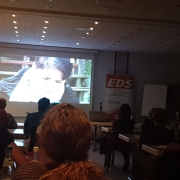 Filmed from the end of a room. In front of the room is a large screen showing 'We Are Visible', Karina's film about Ehlers-Danlos syndrome.