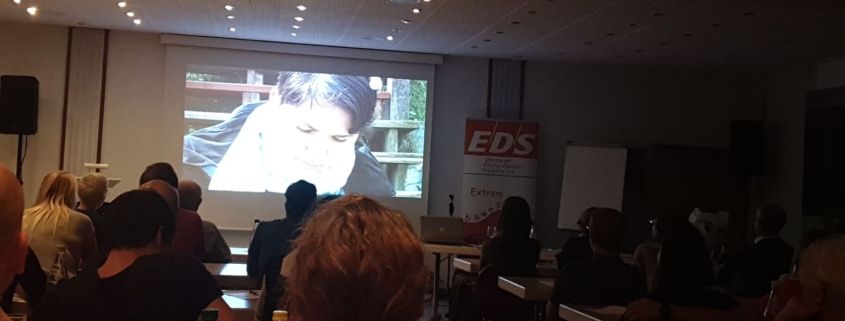 Filmed from the end of a room. In front of the room is a large screen showing 'We Are Visible', Karina's film about Ehlers-Danlos syndrome.