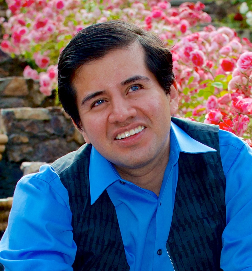 Belo Cipriani: A man with short, brown hair wearing a blue shirt is sitting on stairs with pink flowers in the background. He looks into the distance and is smiling.
