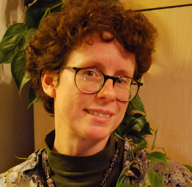 A person with short, brown, curly hair and round, black glasses smiles.