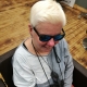 Andrea Eberl, eine Frau mit kurzen, hell-blonden Haaren sitzt auf einem Friseurstuhl und trägt eine schwarze Sonnenbrille