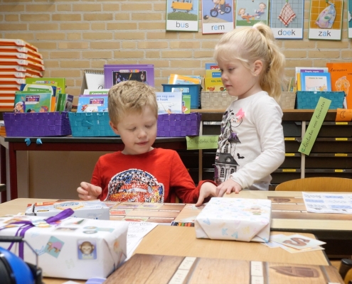 Lucas and Lotte, two young children, are in their classroom playing on a table. Lucas has blonde hair and wears a red "car" shirt, and Lotte has blonde, long hair bound to a ponytail and wears a white shirt.