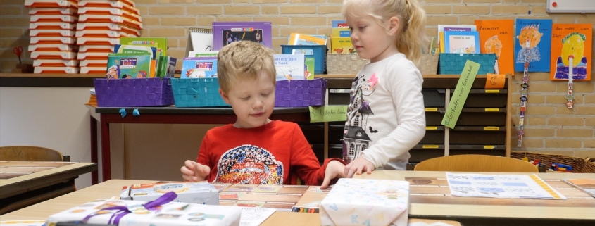 Lucas and Lotte, two young children, are in their classroom playing on a table. Lucas has blonde hair and wears a red "car" shirt, and Lotte has blonde, long hair bound to a ponytail and wears a white shirt.