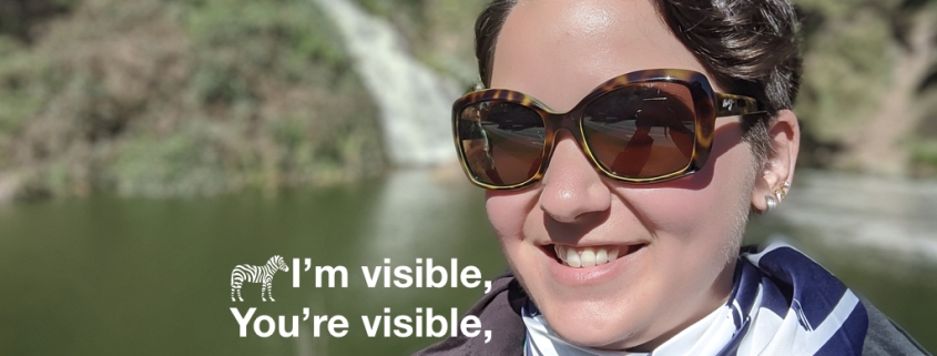 Karina, eine Frau mit kurzen braunen Haaren und einer Blume im Haar lächelt in die Kamera. Sie steht vor einem Wasserfall und trägt ein Zebragestreiftes Halstuch. Auf dem Foto steht: Happy Rare Disease Day 2020, I am Visible, You are Visible, We Are Visible!