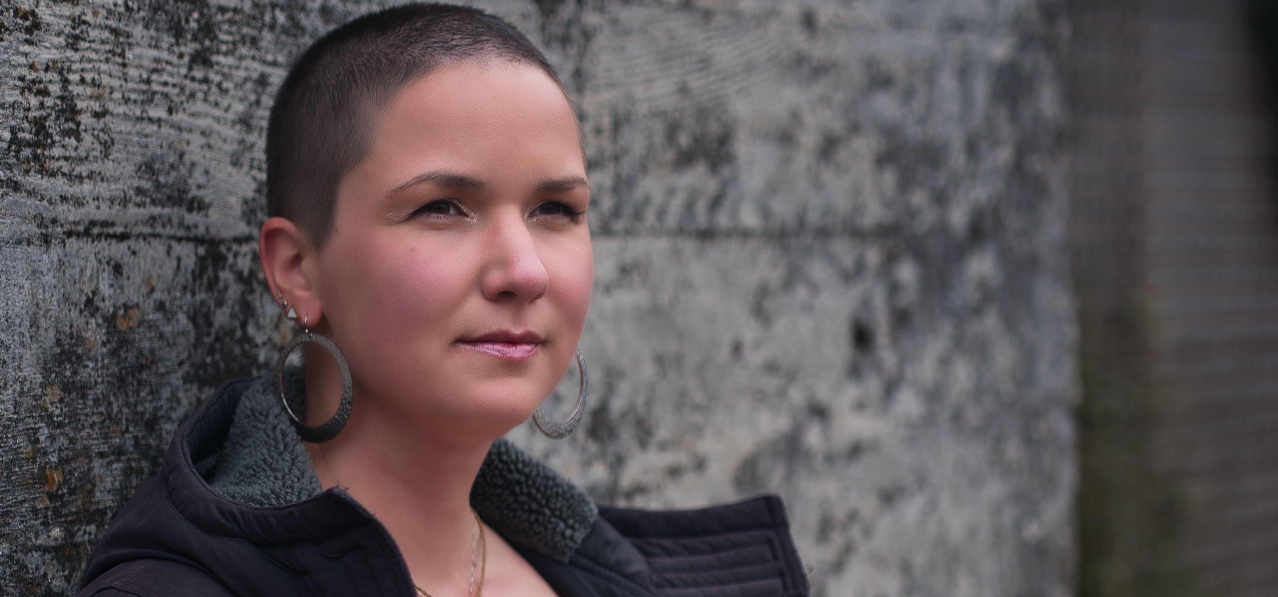 Karina, a woman with shaved, brown hair stands on a grey wall and wears huge earrings.