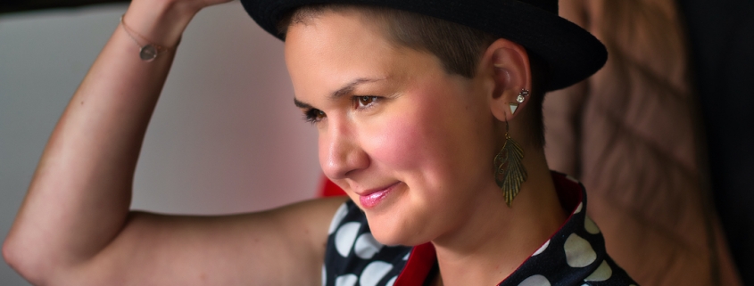 Karina, a woman with super short brown hair is wearing a black hat and has her hand on top of the hat to take it off. She wears huge, feather-like earrings and lipstick.