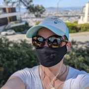 Karina, eine Frau mit schwarzem Mundschutz steht auf einem Hügel mit Aussicht auf die San Francisco Bay. Sie trägt ein T-Shirt, eine Sonnenbrille und eine Türkise Baseball Cap.