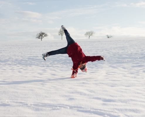 Ein Feld bedeckt von Schnee und wenige kahle Bäume. In der Mitte ist eine Frau, die Winterkleidung trägt und einen einarmigen Handstand macht.