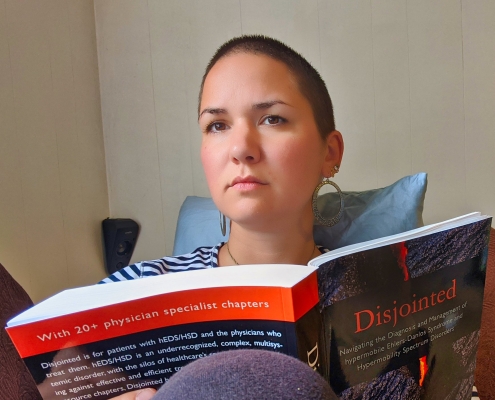 Karina, eine Frau mit super kurzen, braunen Haaren sitzt auf einer Couch und hält das Buch 'Disjointed' in Händen.