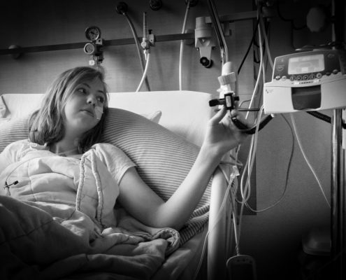 Black and white photo of Jade, a young girl with shoulder-long, blonde hair and a tube up her nose. She faces towards her left hand in which she holds part of her IV