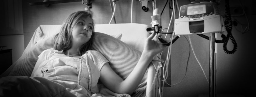 Schwarz-weiß Foto von Jade, einem jungen Mädchen mit blonden, schulterlangen Haaren in einem Krankenhausbett. Sie hat einen Schlauch in der Nase und blickt zu ihrer linken Hand, in der sie einen Teil ihrer Infusion hält.