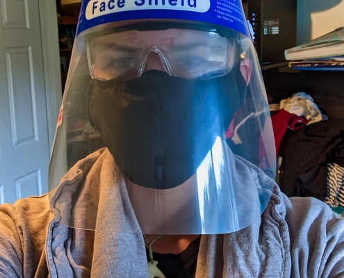 Karina, eine Frau mit super kurzen braunen Haaren trägt ein Face Shield, eine Laborbrille und drei Masken übereinander.