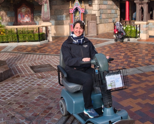 Karina, eine Frau mit kurzen, braunen Haaren sitzt in einem elektronischen Scooter in Disneyland vor bunten Häusern.