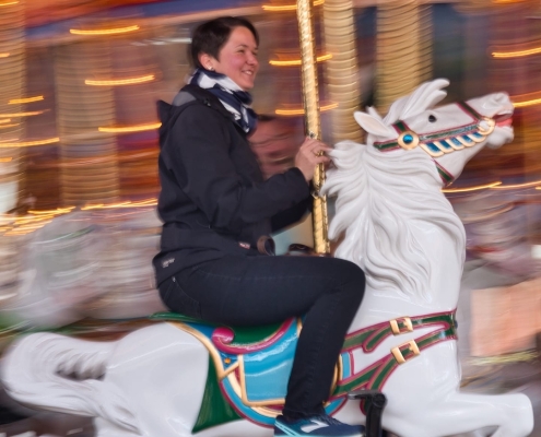 A woman with short brown hair sits on a white carousel horse and smiles.