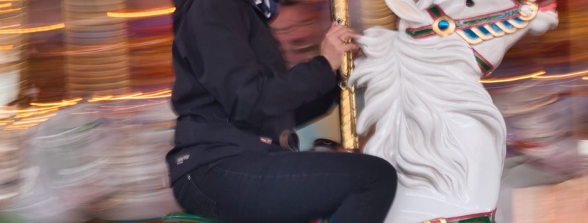 A woman with short brown hair sits on a white carousel horse and smiles.