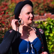 Karina, a woman with shaved head, is standing in her backyard wearing a black hat, red lip stick and large silver hoop earrings