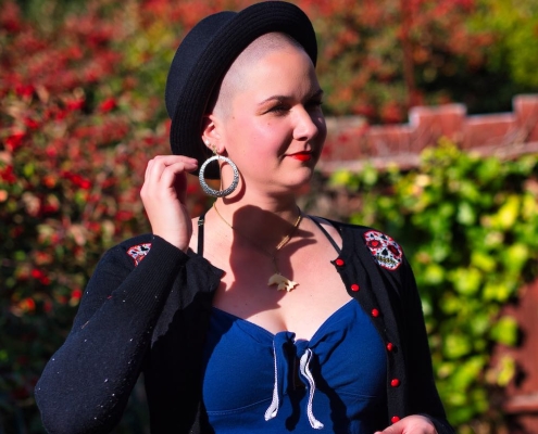 Karina, eine Frau mit abrasierten Haaren, steht in ihrem Garten. Sie trägt einen schwarzen Hut, große silberne Ohrringe und roten Lippenstift.