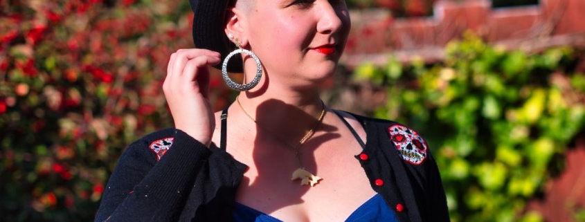 Karina, a woman with shaved head, is standing in her backyard wearing a black hat, red lip stick and large silver hoop earrings