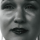 Black and white image of Karina, a woman with shaved head. She is wearing glitter eye shadow and red lipstick.