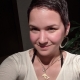 Karina, a woman with short brown hair and a white shirt. She wears large silver hoop earrings and a white dolphin necklace.
