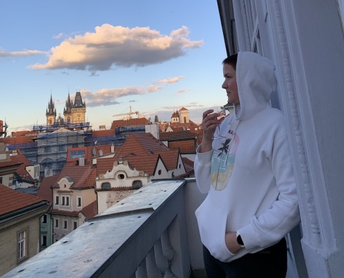 Karina, a woman with short brown hair leans against a wall on a balcony during sunset. She wears a white hoodie and holds a glass of wine in her hand. In the background you can see an old church and many older houses.