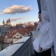 Karina, a woman with short brown hair leans against a wall on a balcony during sunset. She wears a white hoodie and holds a glass of wine in her hand. In the background you can see an old church and many older houses.
