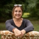 A woman with long brown hair bound to a ponytail sits in her garden on a chair with giraffe pattern. She smiles, wears a black shirt and sunglasses on top of her head.