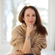 A woman with long brown hair hanging over her left shoulder sits on a chair with her right arm under her chin. She wears a brown sweater and sits in a bright white room.